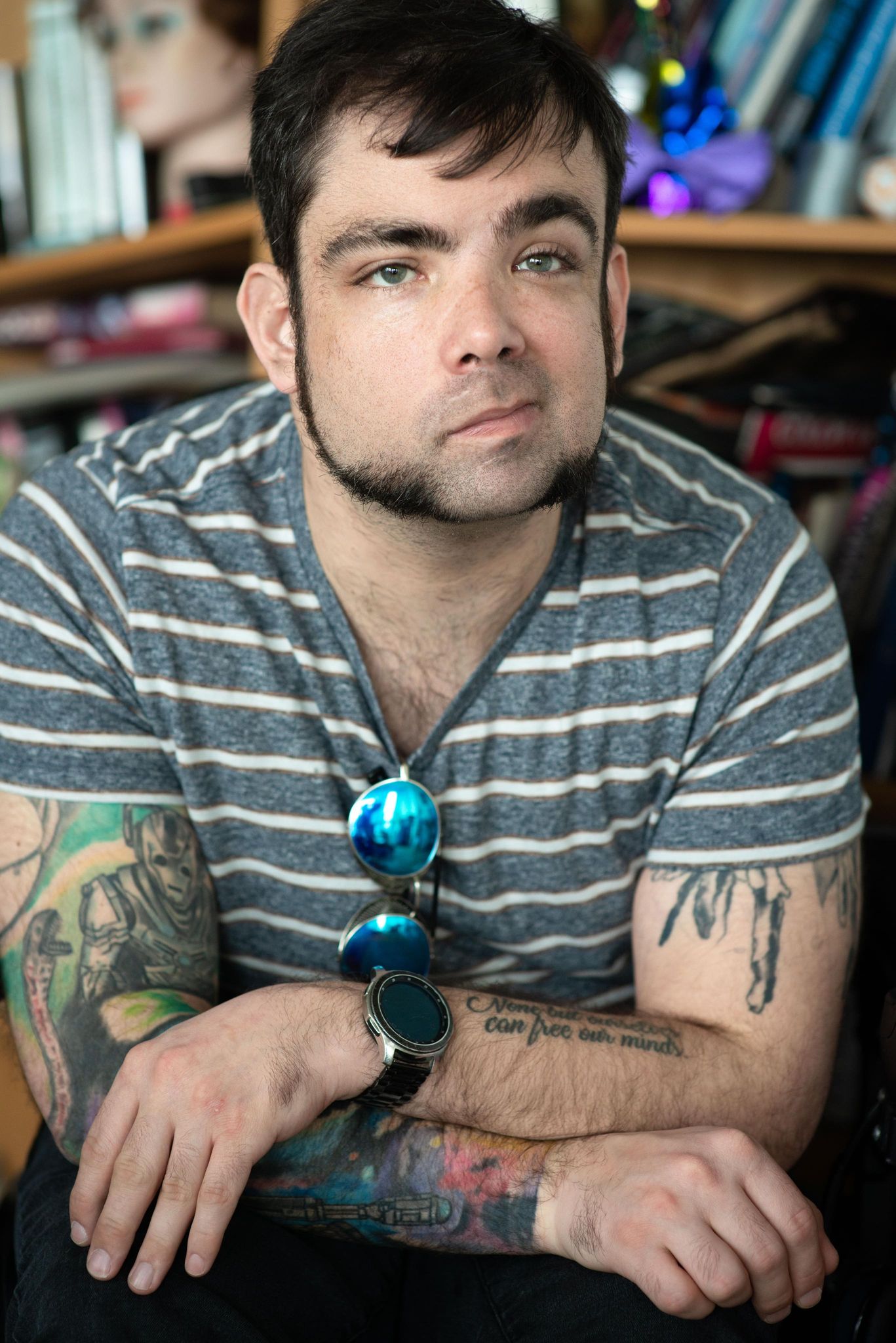 The picture shows a man sitting casually, wearing a striped shirt and sporting unique facial hair. His arms are resting on his lap, displaying several intricate tattoos. He has a pair of reflective sunglasses hanging from his shirt, and he is also wearing a watch on his left wrist. The man’s expression is calm, with a slight gaze forward. The background appears to be indoors, with books and various items visible behind him, suggesting a personal or creative environment. The tattoos on his arms are detailed and cover a significant portion of his skin.