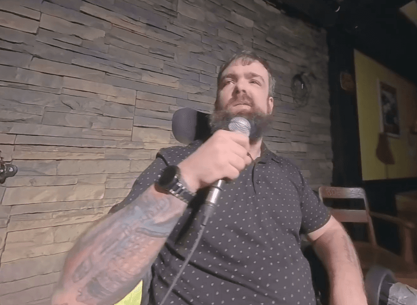 A bearded man seated indoors holds a microphone near his mouth. He wears a dark short-sleeve polo shirt with small dots, a wristwatch, and has tattoos on his forearm. A stone-textured wall and wooden chair are visible in the background.