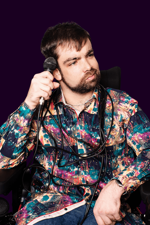 A man seated in a wheelchair holds a microphone beside his head. He has short dark hair and a beard, and wears a brightly patterned multicolor button-up shirt. Black microphone cables are draped across his chest against a dark purple background.
