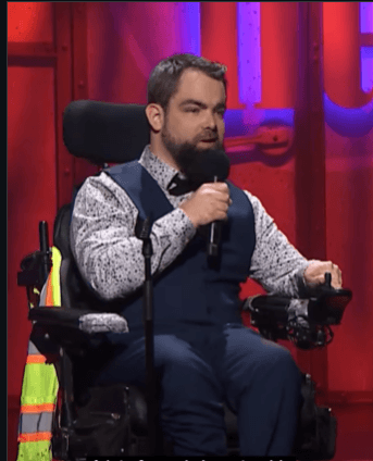 A man seated in a powered wheelchair speaks into a handheld microphone on a stage with red and purple lighting. He wears a patterned white shirt, blue vest, bow tie, and matching pants. A high-visibility vest hangs on the side of the wheelchair.