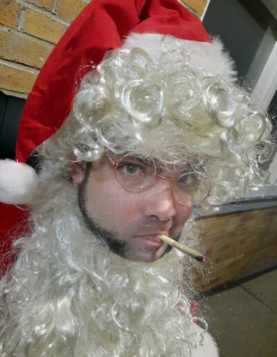 This image shows a person dressed as Santa Claus with a humorous twist. They are wearing the iconic red Santa hat and have a curly white wig and beard. The person is also wearing round glasses and is smoking a lit joint, giving the traditional Santa a rebellious and laid-back vibe. The background has a brick wall and what looks like an outdoor setting. The overall image blends the festive holiday look with a bit of humor and edge.